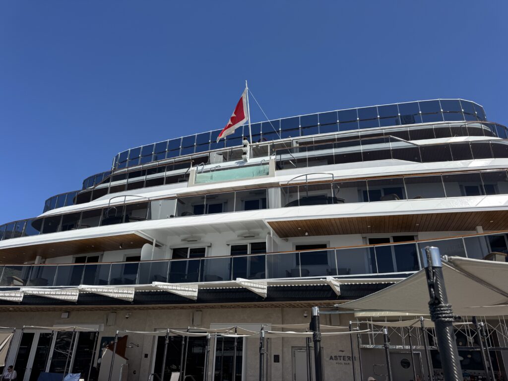 Top deck of a modern luxury yacht with glass railings, a red-and-white flag, and a clear blue sky.