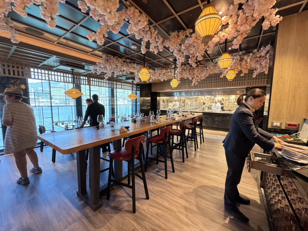 Open kitchen restaurant with a long wooden table, pink blossom garlands, and yellow lanterns.