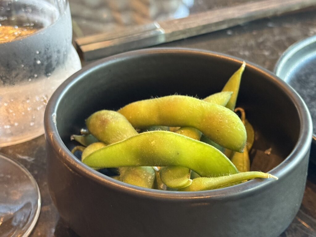 Fresh green beans in a dark bowl on a wooden table, with a glass jar nearby.
