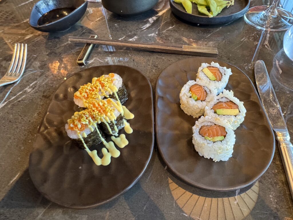 Assorted sushi rolls on dark plates with salmon and avocado fillings, drizzled with sauce on the left plate and chopsticks nearby on a marble table.