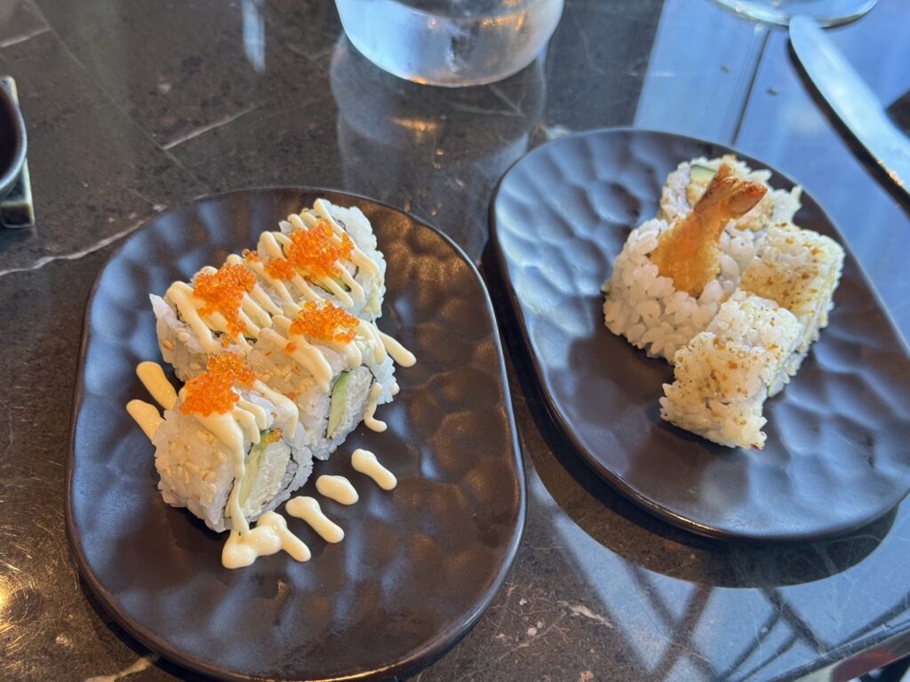 Sushi rolls on dark plate topped with orange tobiko and drizzled mayonnaise, with another plate of fried shrimp-temaki style sushi nearby.