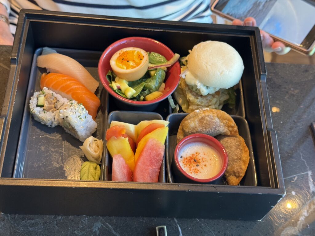 Assorted sushi and side dishes in a black bento box, including salmon nigiri, cucumber roll, salad, a boiled egg cup, and dipping sauces.