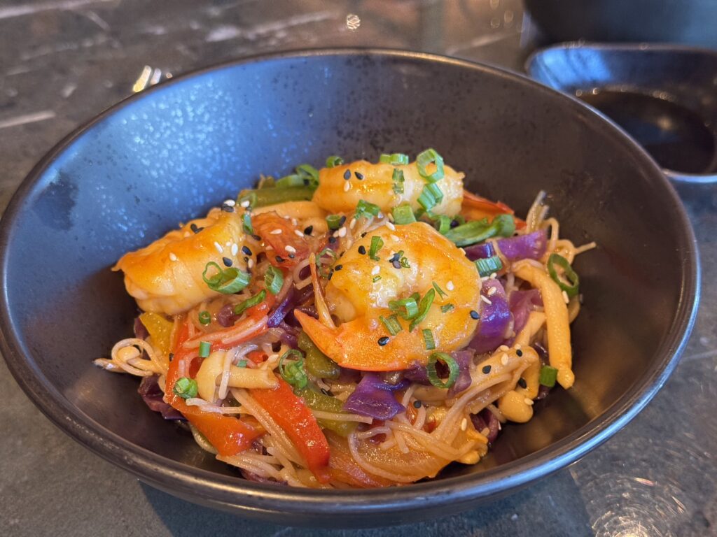 Bowl of colorful shrimp fried noodles topped with sesame seeds and chopped green onions, in a dark ceramic bowl.