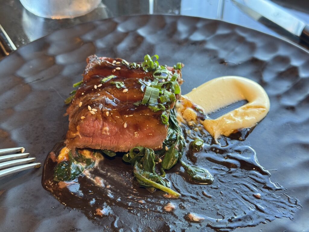 Beef slice with glossy glaze, sesame seeds, and chopped green onions on a bed of greens with a lemon wedge nearby on a dark plate.