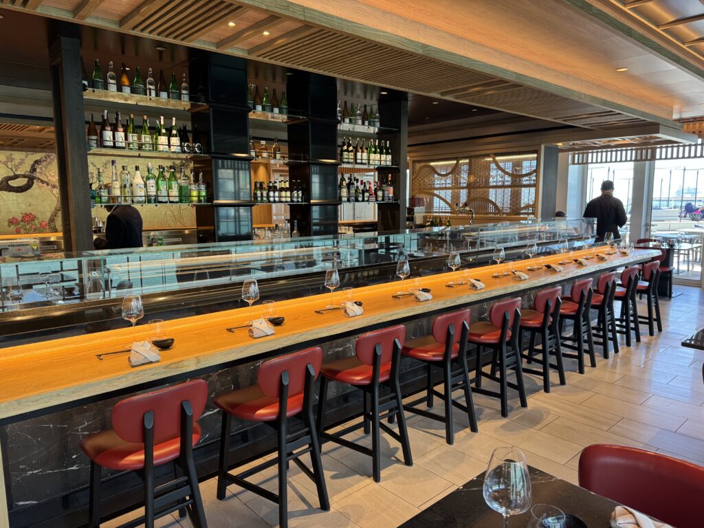 Long bar with a marble-and-wood counter, red high stools, and shelves stocked with bottles behind a glass barrier in a modern restaurant.
