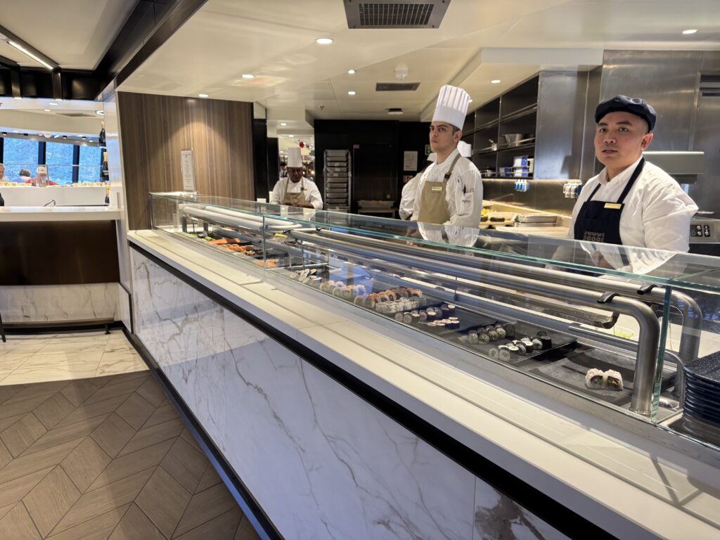 Sushi display case with various nigiri rolls; three chefs in white uniforms and tall hats stand behind the counter behind glass.
