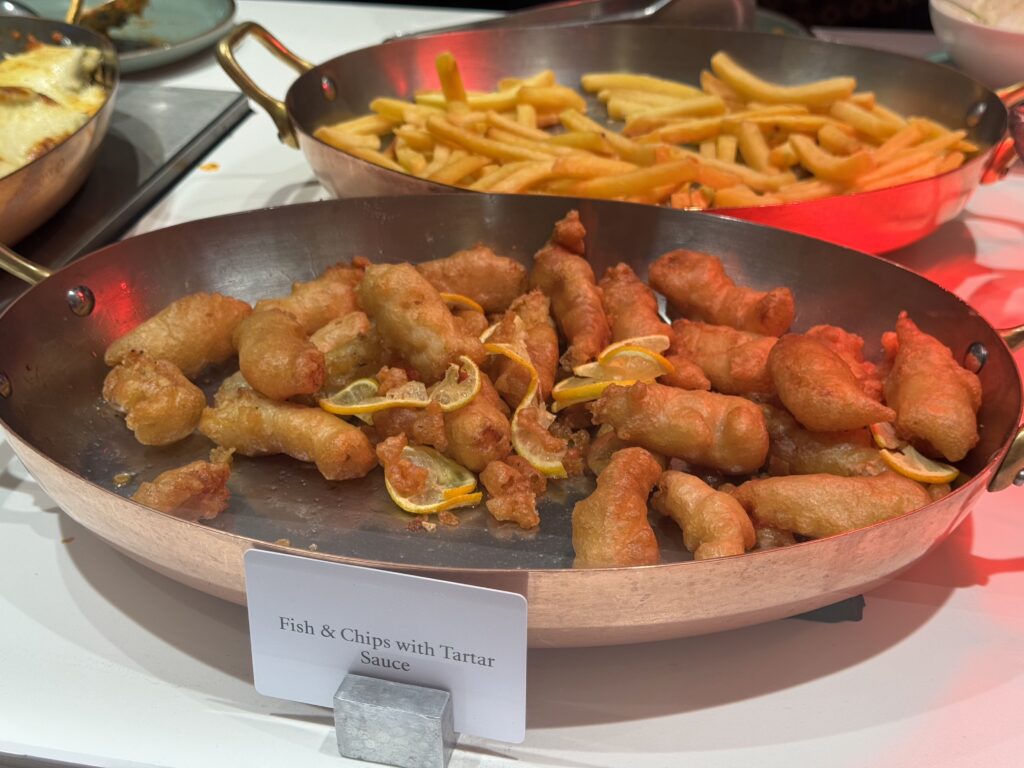 Golden fried fish bites with lemon wedges in a shallow pan, with a separate pan of french fries in the background.