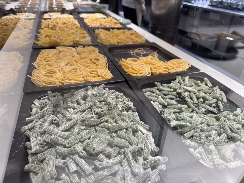 Assorted trays of pasta on display, including green spinach pasta and plain noodles in a deli counter