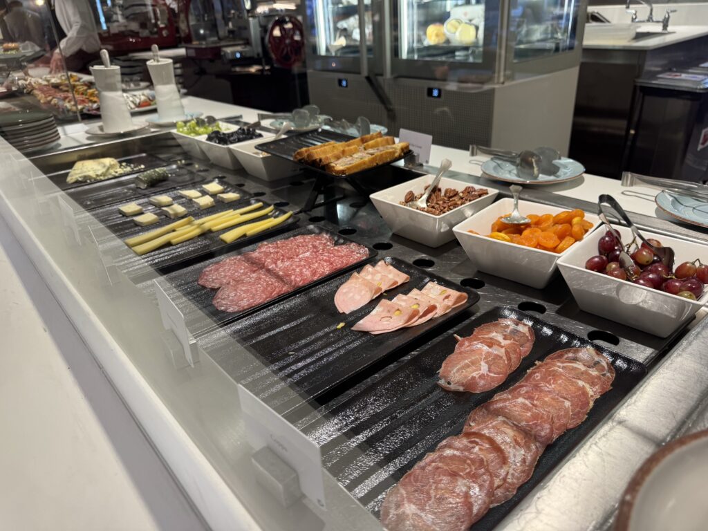 Deli counter with sliced meats, cheese cubes, bread slices, olives, nuts, and bowls of apricots and grapes on display.