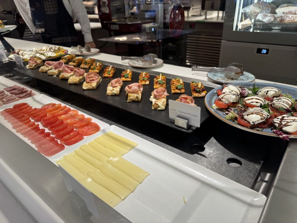 Assorted bite-sized canapés and skewers arranged on a black serving tray at a deli counter, with tomatoes and cheese slices in foreground and glass display in background