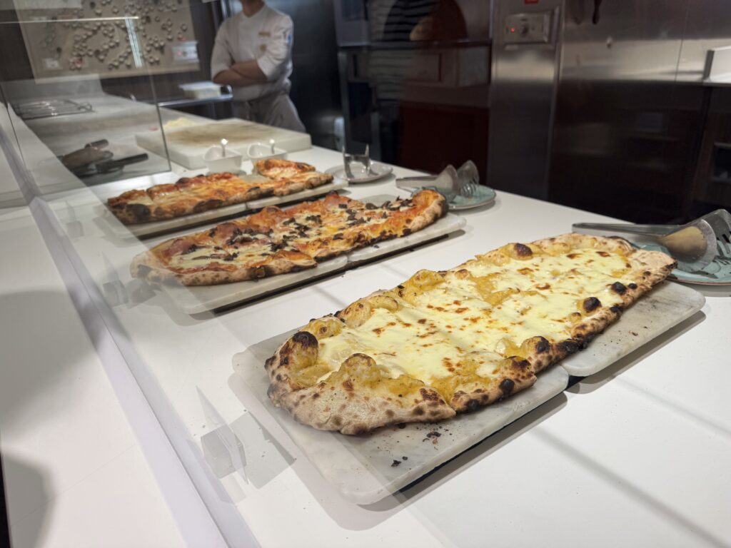 Pizzas on trays inside a glass display, cheese and toppings visible, in a commercial kitchen setting