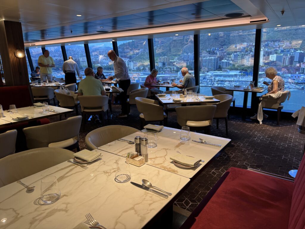 Restaurant dining area aboard a cruise ship with panoramic windows and a harbor view; guests are seated at marble tables.