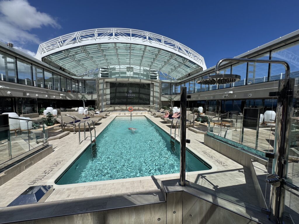 Outdoor hotel/cruise pool deck with glass railings, loungers, and a modern glass-wabled atrium under a blue sky, front view of the pool area.