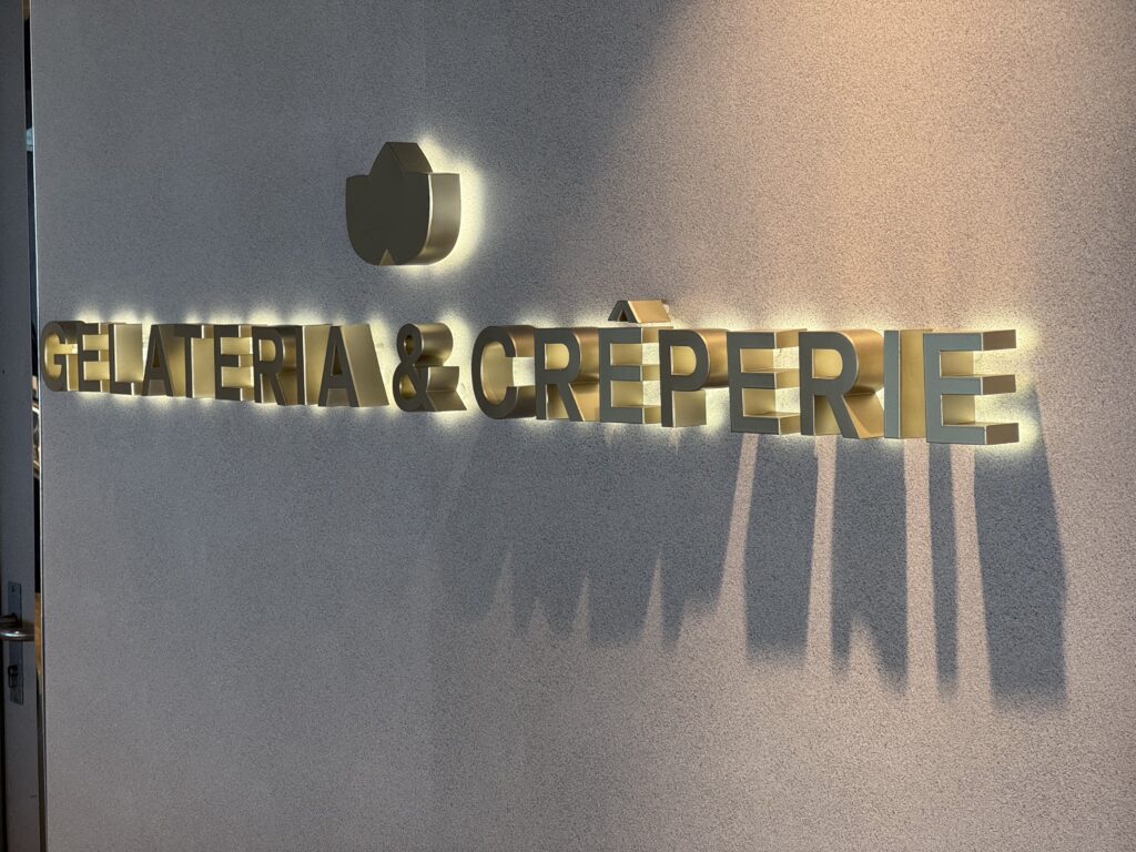 Gold 3D sign reading 'GELATERIA & CREPERIE' on a textured wall with a round logo above.