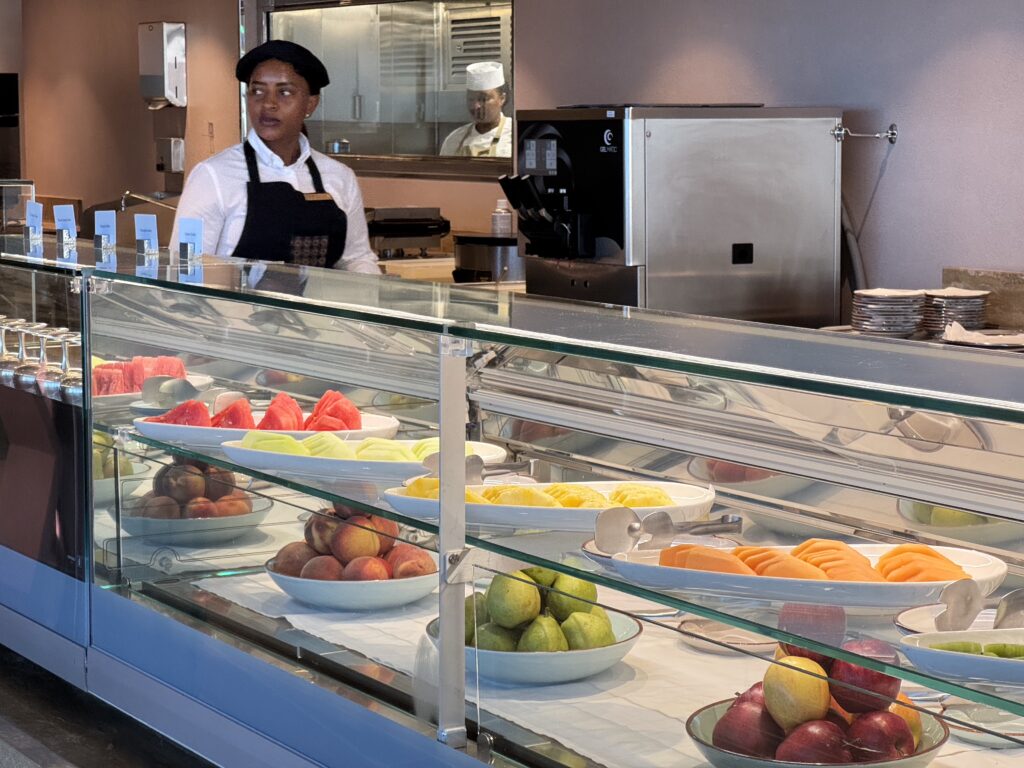 Fruit display counter with bowls of sliced fruit (watermelon, melon, pineapple) behind glass; two staff members in white shirts and aprons working in the kitchen.
