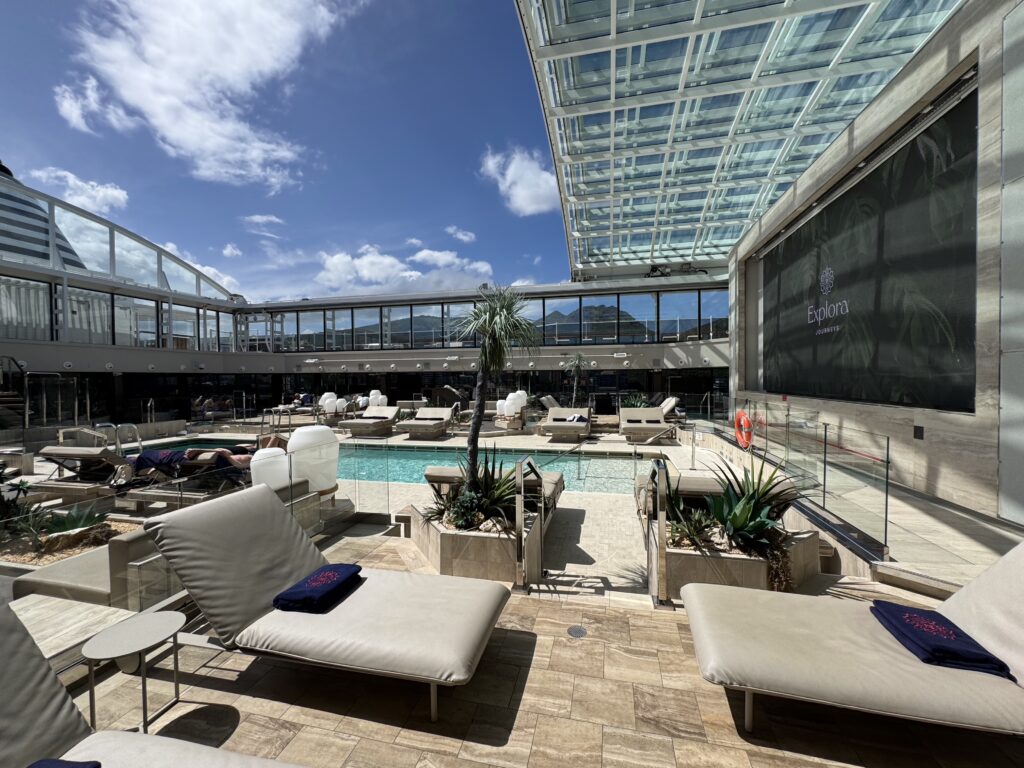 Outdoor pool deck with beige lounge chairs, glass railing, and tropical plants under a bright blue sky at a modern resort.