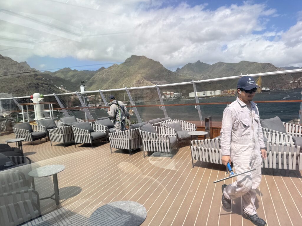 Outdoor yacht deck with wicker seating and glass railing; man in cap and sunglasses stands to the right with mountains and water in the background.