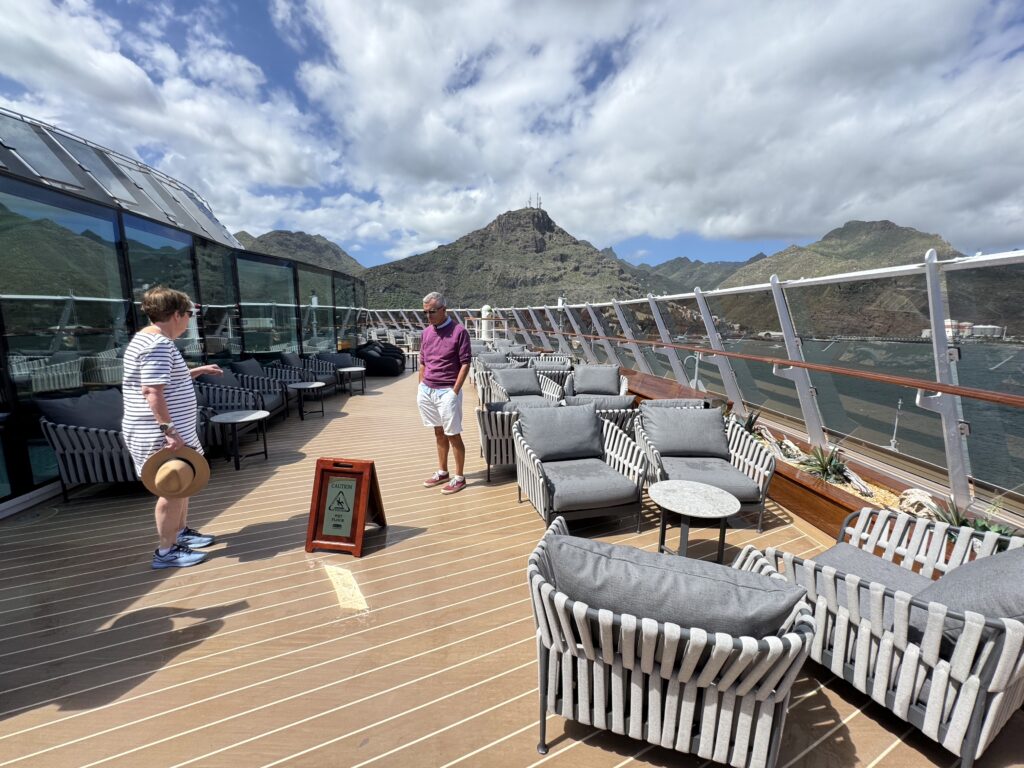 People stand on a rooftop lounge deck with rows of gray cushioned chairs, glass railing, and mountain scenery in the background under a partly cloudy sky, a man in a purple shirt and another in a striped shirt nearby.