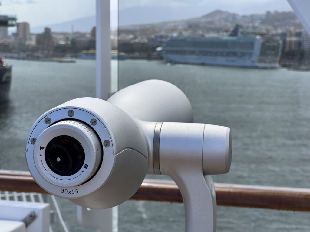 Close-up of a metallic security camera mounted on a railing, with a harbor and distant cruise ship blurred in the background.