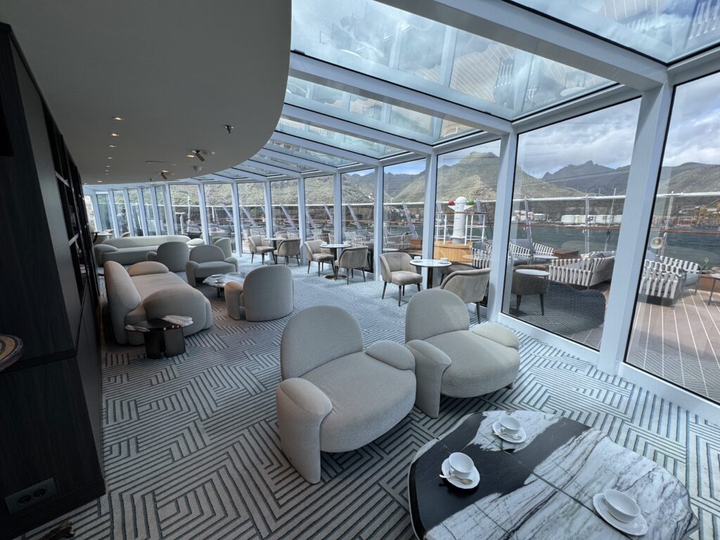 Lounge area inside a glass-walled deck with light gray curved sofas, round tables, and patterned carpet, overlooking mountains and water outside.