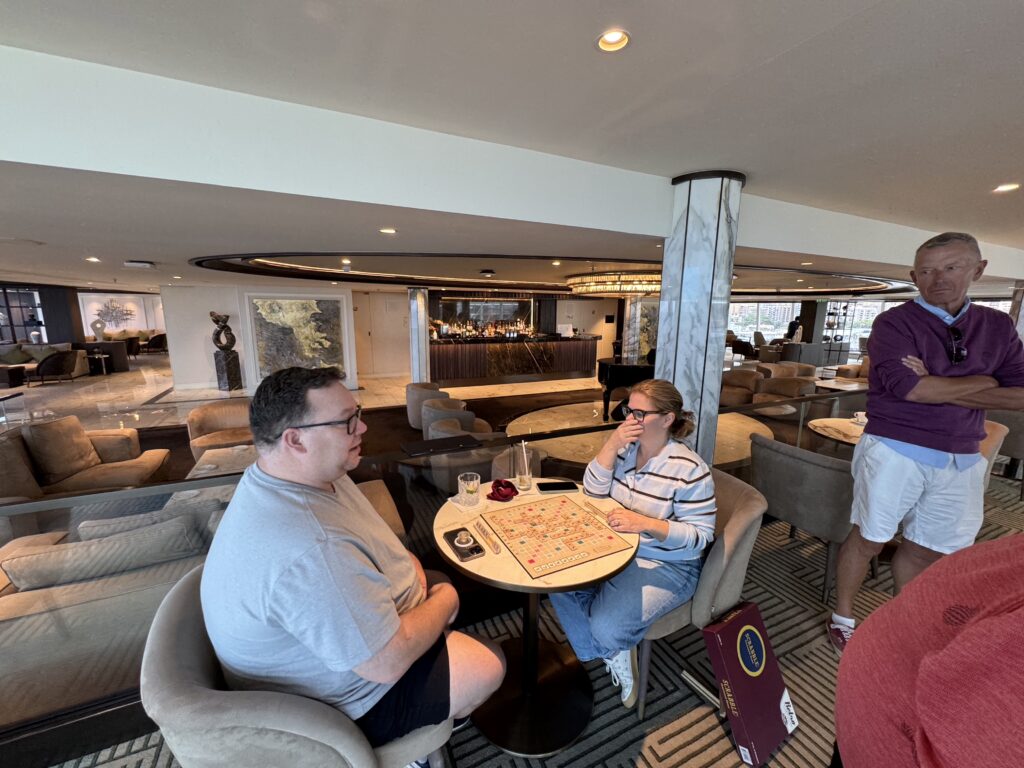 Three people sit around a circular Scrabble table in a hotel lounge, playing the game while a man stands nearby by a marble pillar.