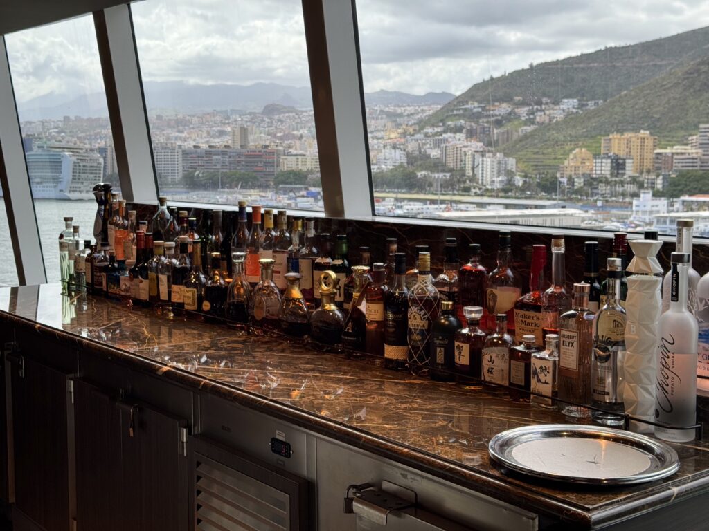 Bar counter filled with assorted liquor bottles, marble surface, large windows overlooking a city and hills outside
