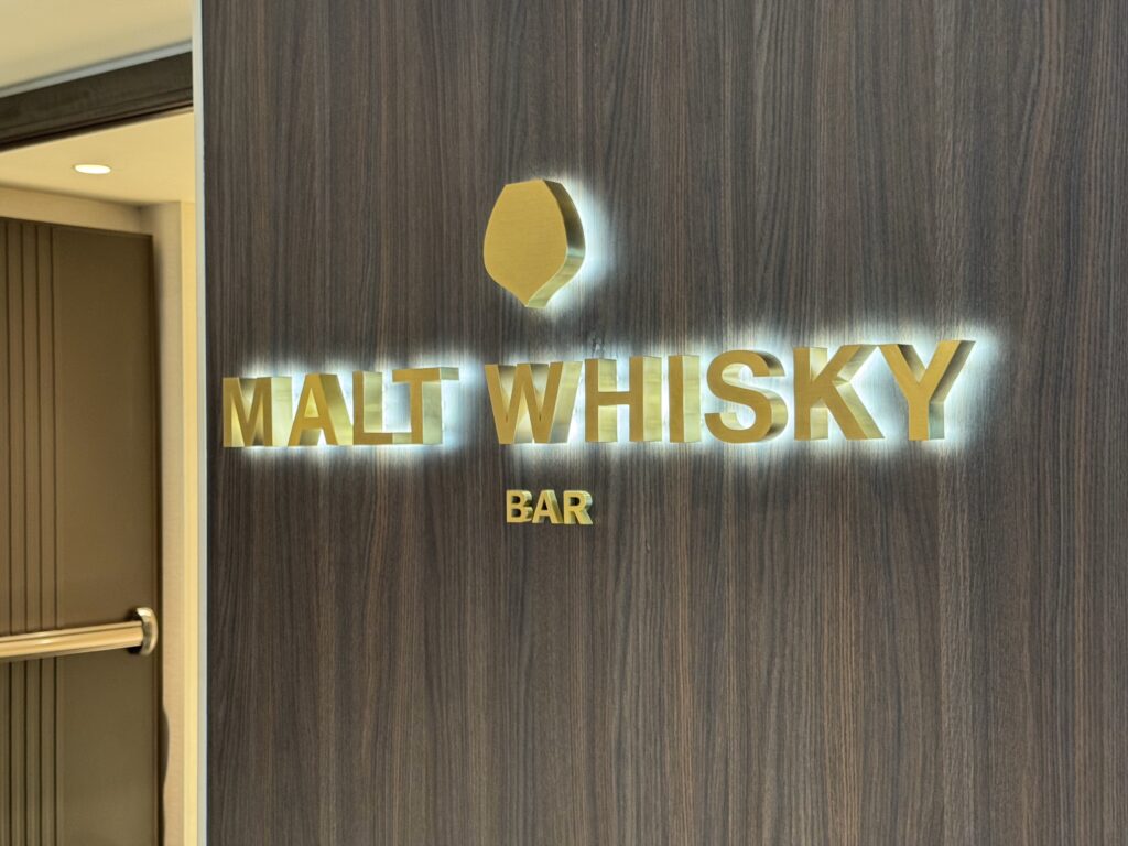 Sign on dark wood wall reading 'MALT WHISKY' with a gold circular logo above, and 'BAR' visible below.