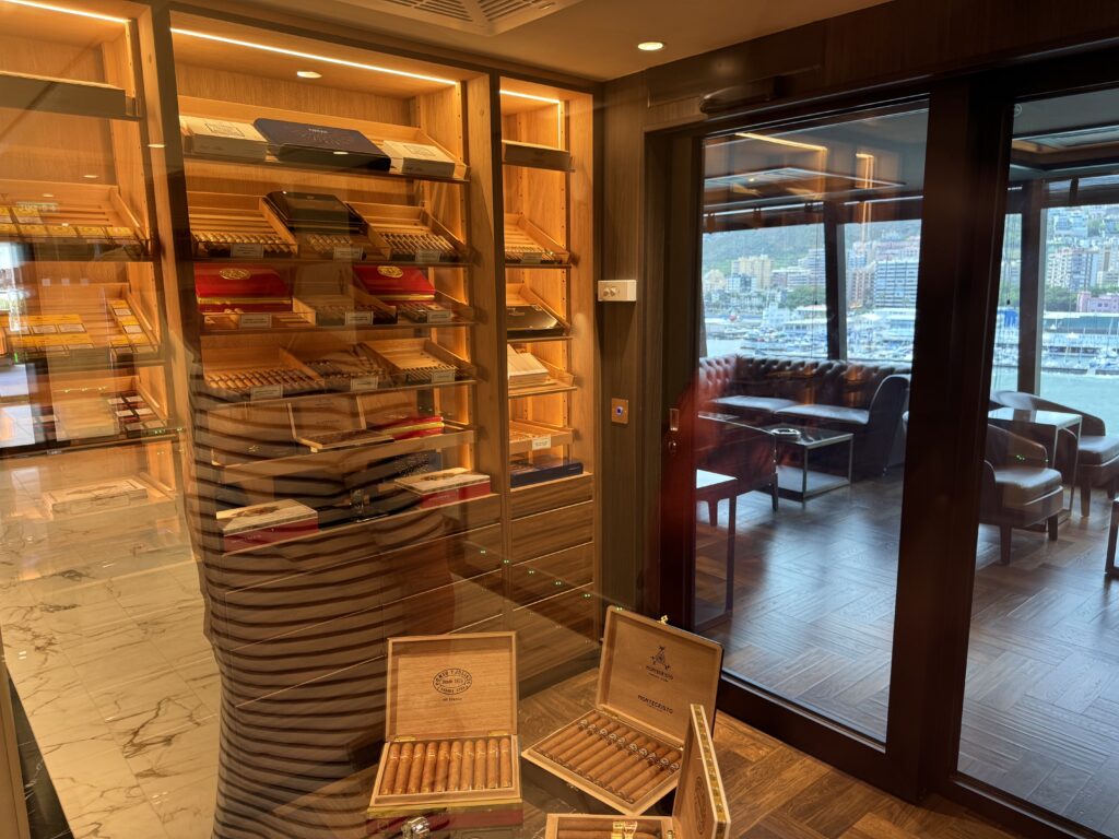Wooden cigar display cases with open boxes of cigars, glass doors reflecting the showroom, and a harbor view through large windows.