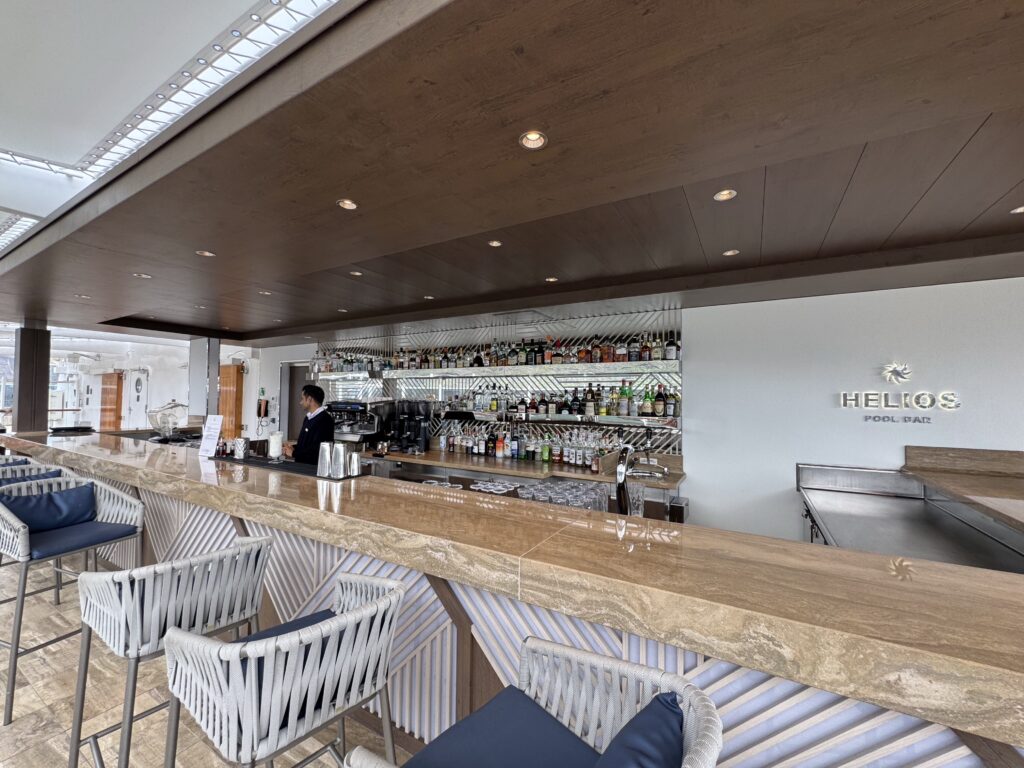 Modern pool bar interior with a wood-paneled ceiling, recessed lights, and a long marble counter lined with striped stools.