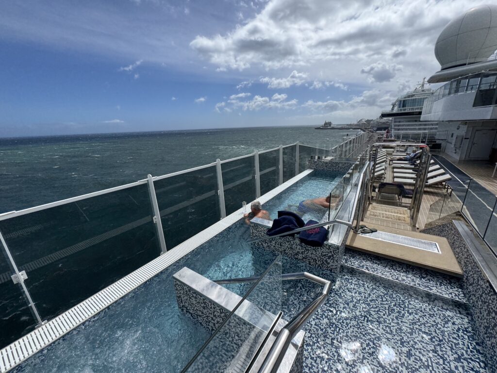 Cruise ship pool deck with glass railing, loungers, and the ocean beyond under a partly cloudy sky