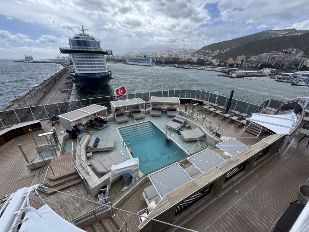 Large cruise ship docked at a port, viewed from a pool deck with lounge chairs and glass railing, city and hills in the background.