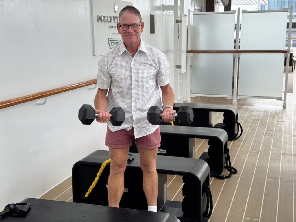 Smiling man in a patterned shirt holds dumbbells while standing on a treadmill in a bright gym.
