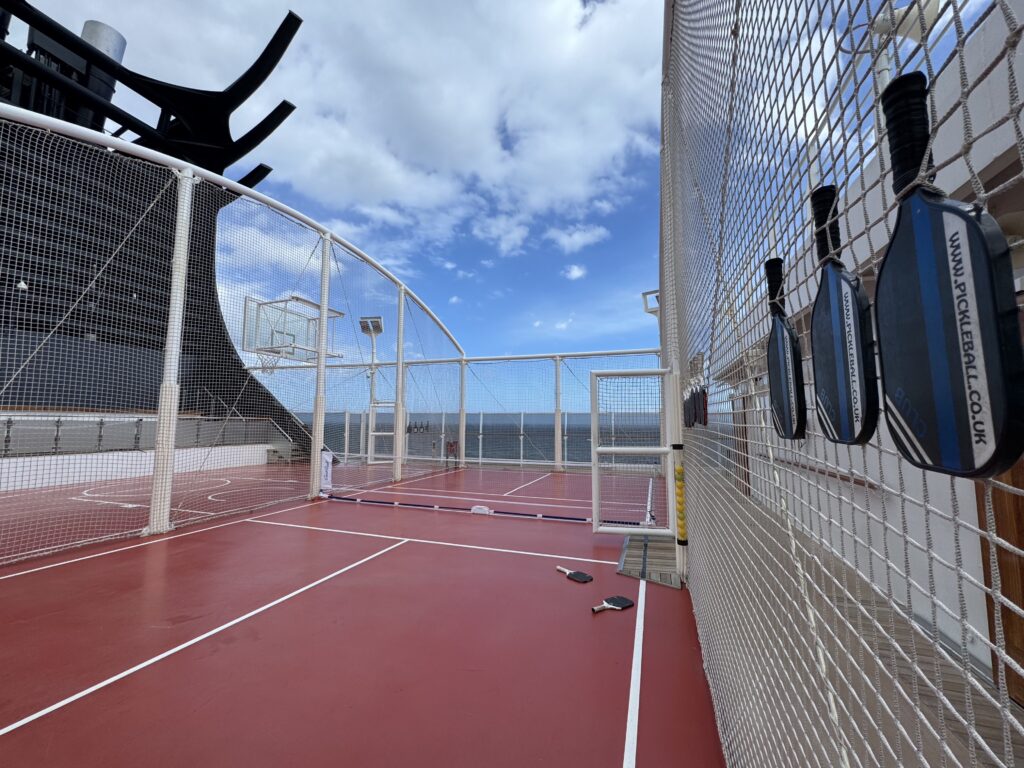 Open-air padel court with a red surface, white boundary lines, and blue paddles hung on the surrounding fence.