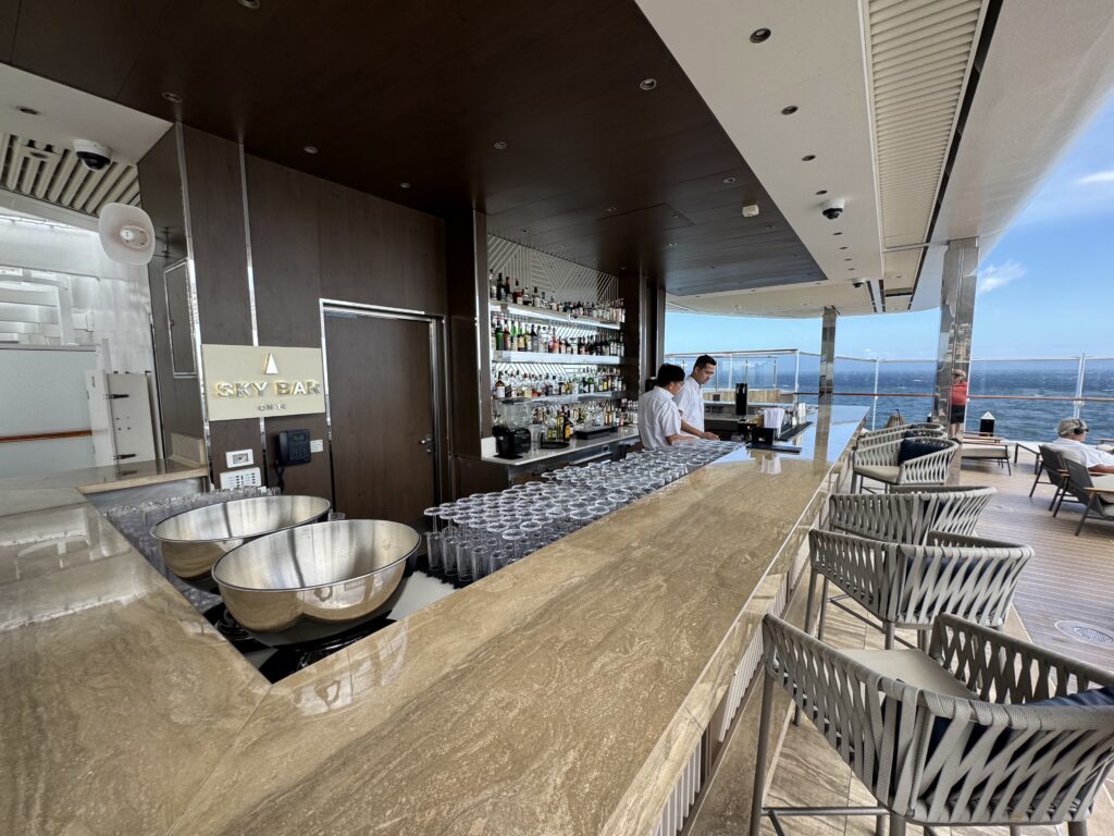 Open-air cruise ship sky bar with a marble counter, liquor bottles on shelves, and glassware, overlooking the ocean.