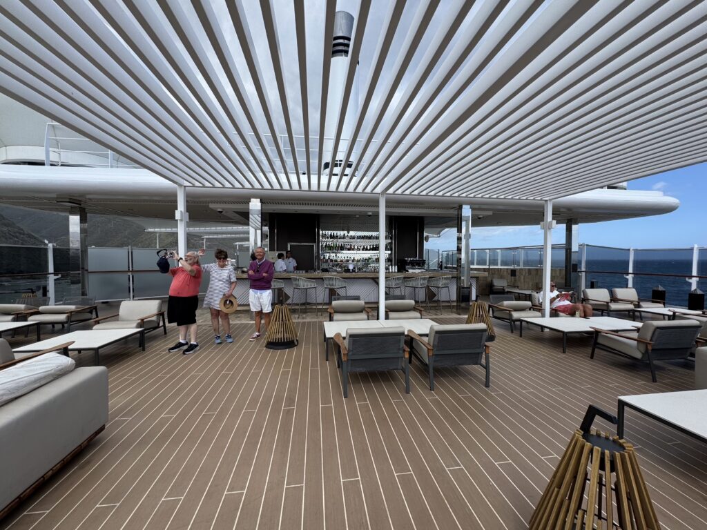 Open-air rooftop lounge with a white slatted canopy, bar in the back, and people standing on a wood-decked terrace overlooking the water.