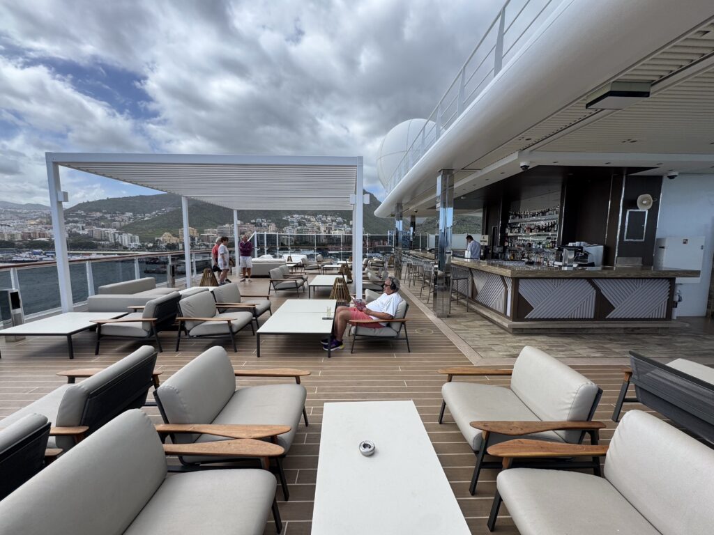 Open-air cruise deck with white lounge chairs, tables, and a bar, overlooking a city and hills under a cloudy sky.
