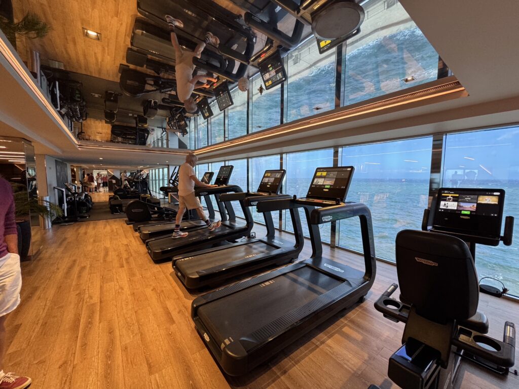 People using treadmills in a spacious cruise ship gym with ocean view through large windows, beside wooden floors.