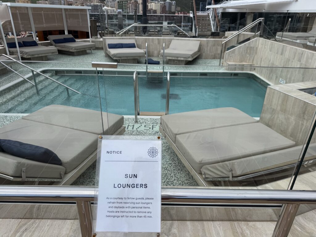 Cruise ship pool deck with beige cushioned loungers along glass railing and city skyline beyond; foreground sign reads NOTICE SUN LOUNGERS.