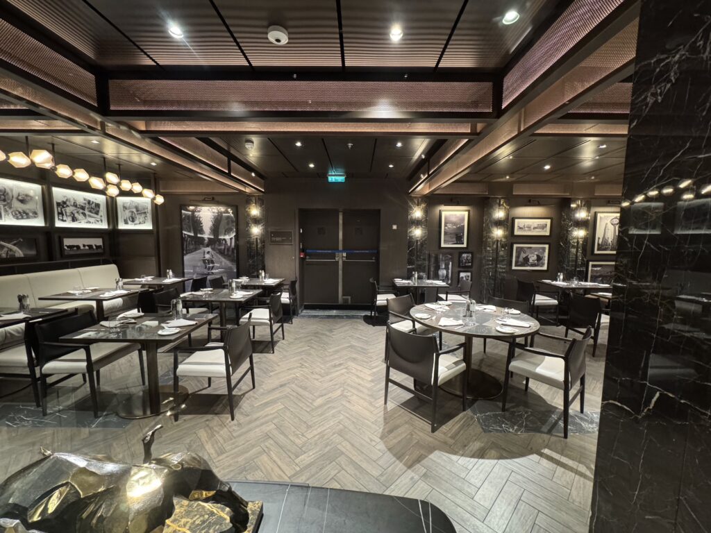 Upscale restaurant dining area with multiple tables and chairs, wall art, and warm lighting in a dark, modern interior.