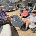 Three adults sit around a dark-marble table on a sunny cruise-ship deck, each with a tropical cocktail.