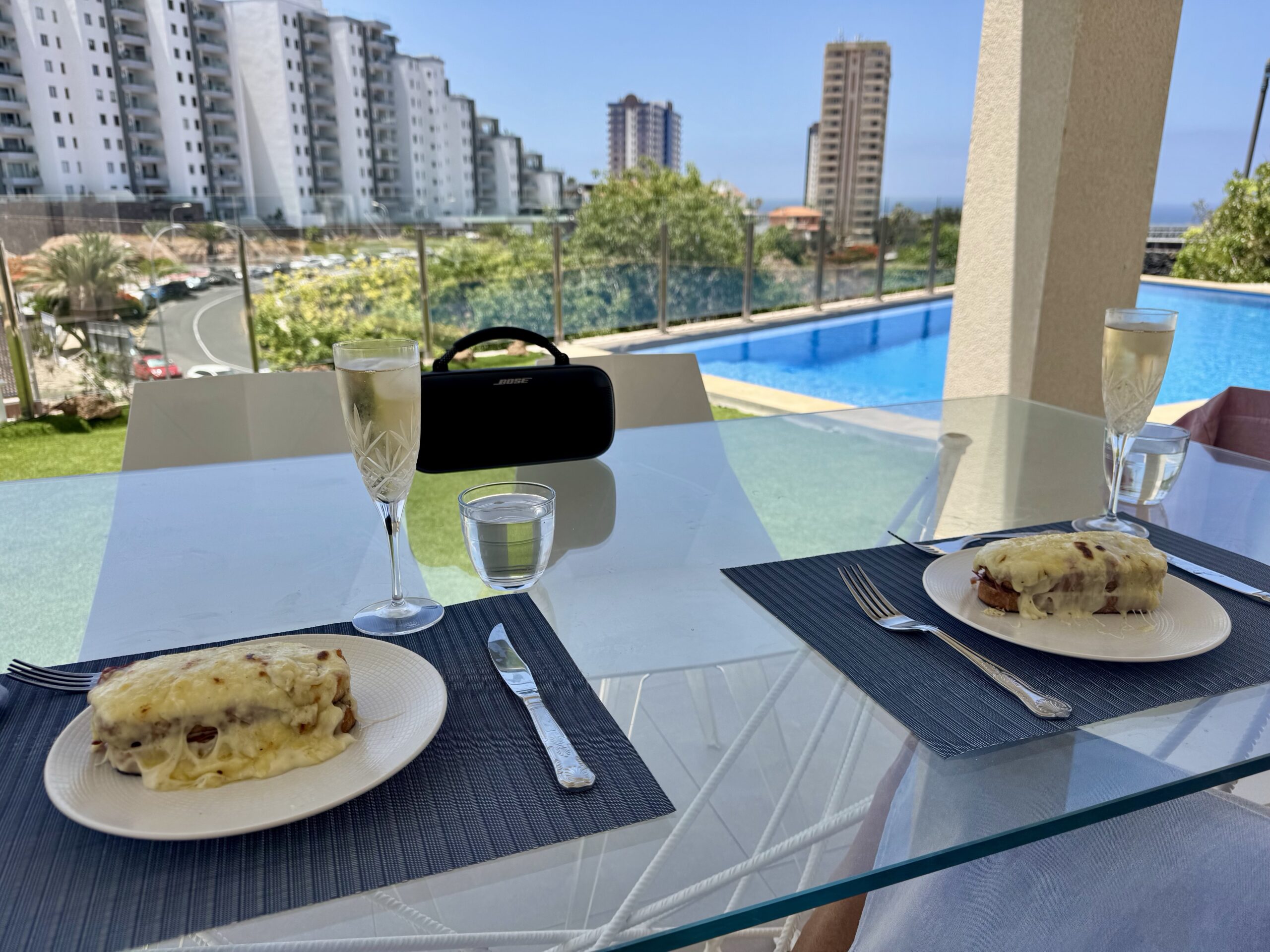 Poolside dining scene with pastries and drinks on a glass table, a Bose speaker, and a city skyline in the background.