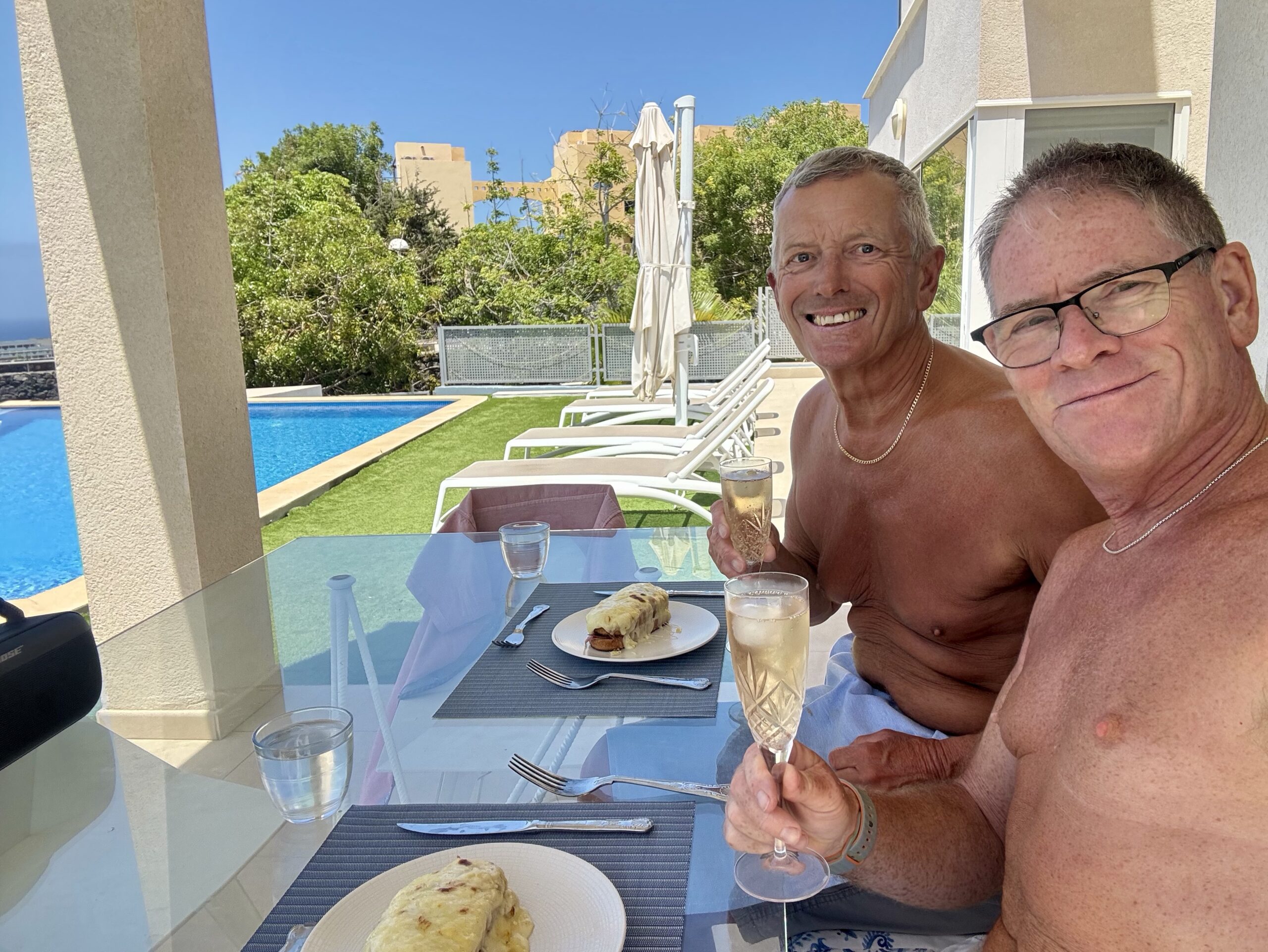 Two men shirtless at a poolside terrace smiling and holding champagne glasses over a glass table with plates of bread/pastry nearby