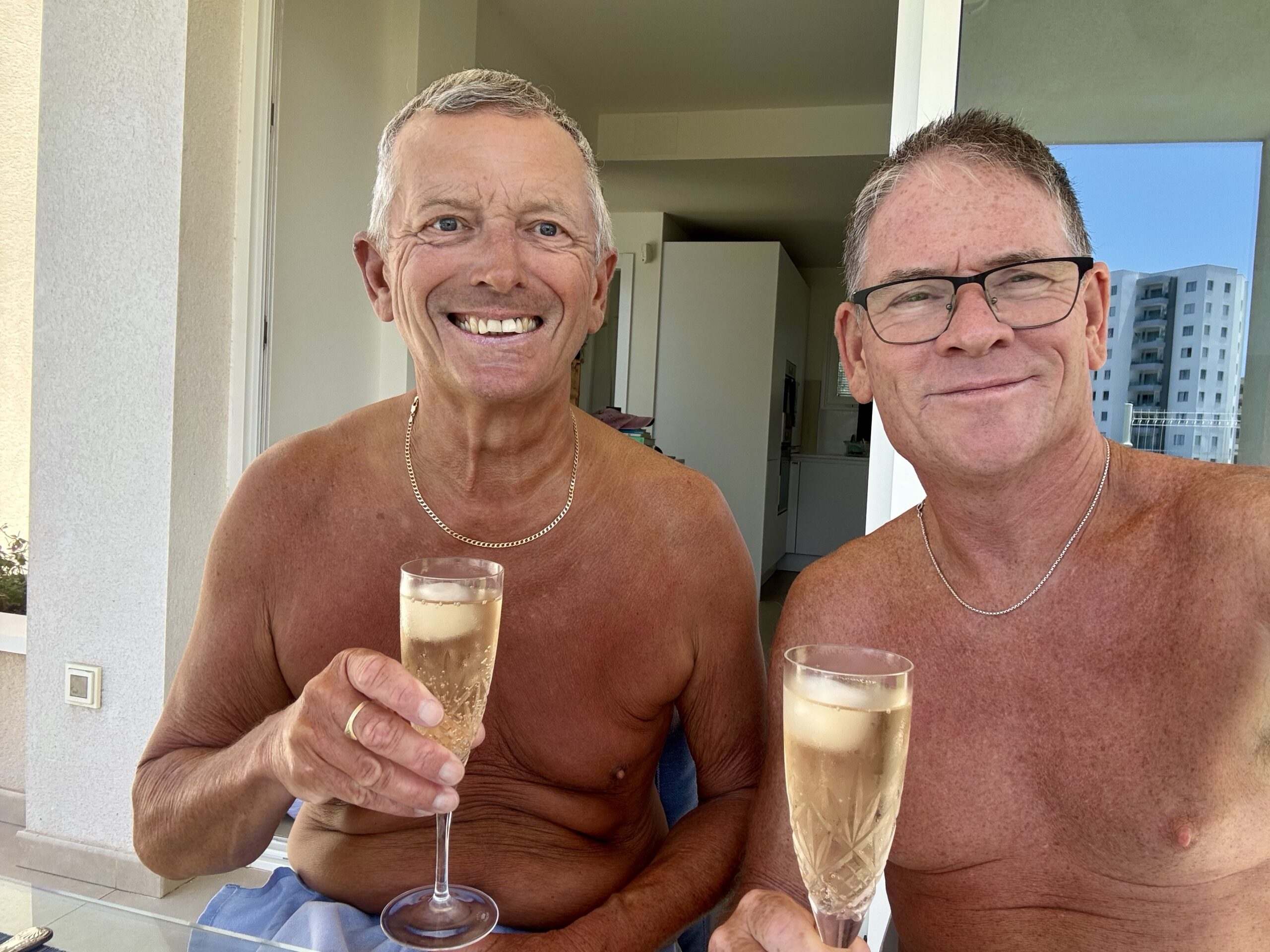 Two shirtless men smiling and toasting with champagne flutes on a balcony, with buildings outside.