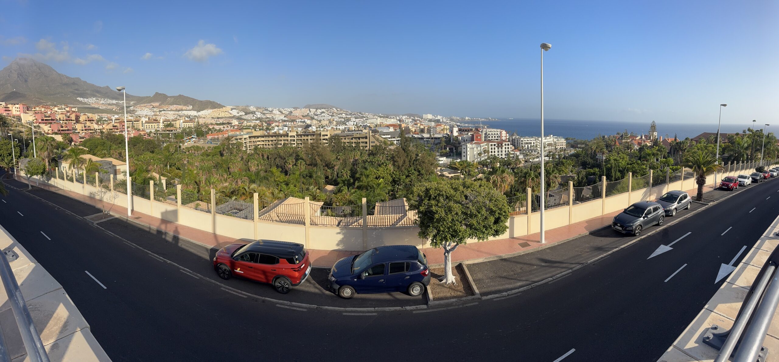 Panoramic view of a coastal city: mountains on the left, numerous buildings in the center, and the sea on the right, seen from a street with parked cars in the foreground.