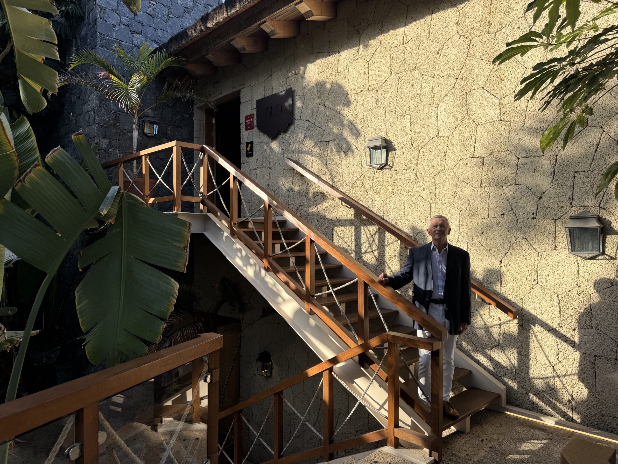 Man in a suit standing on a wooden staircase outside a stone wall building with tropical plants nearby.