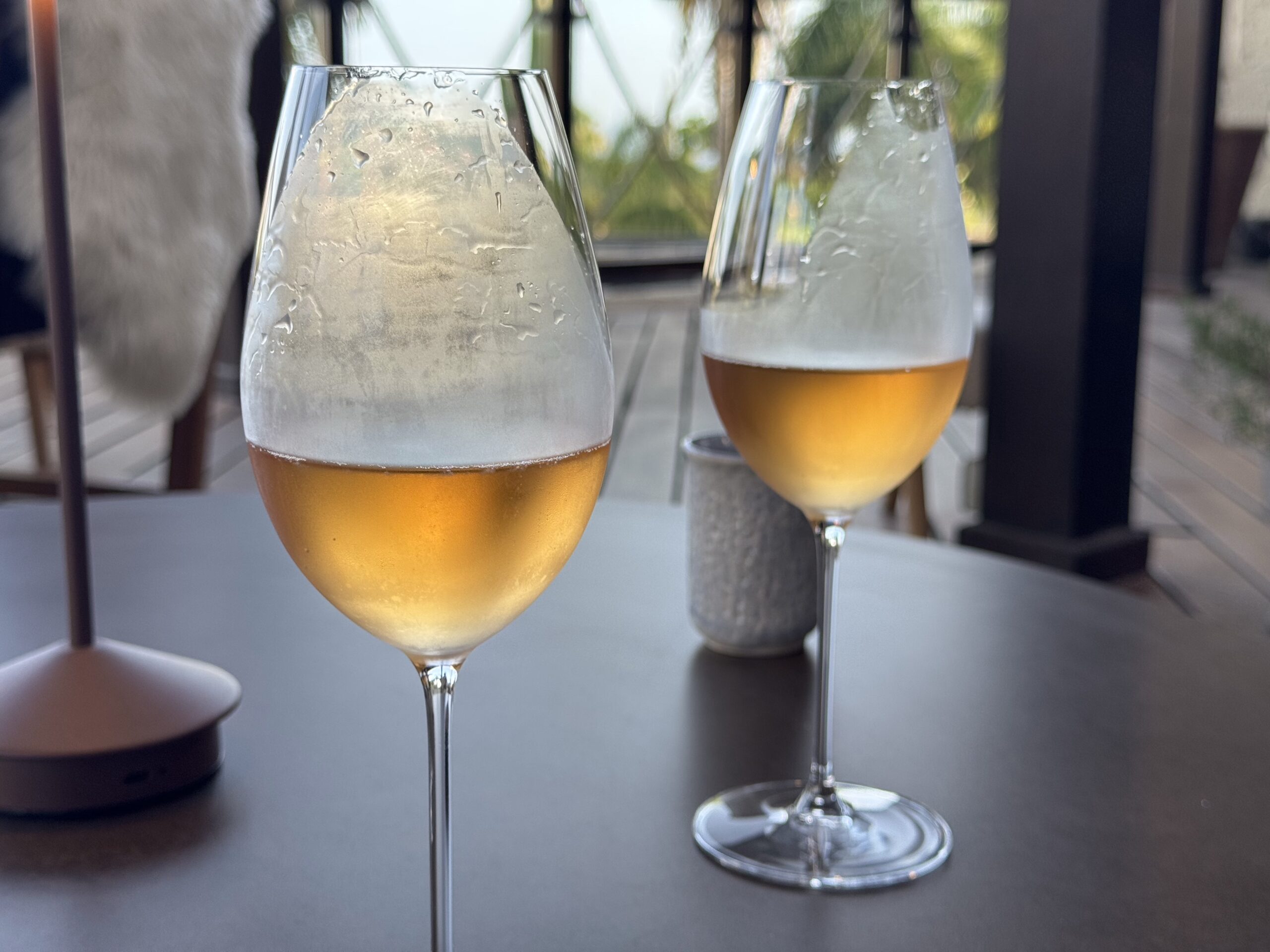 Two beer glasses with foamy tops sit on a dark table outdoors, condensation beads gleaming in daylight.
