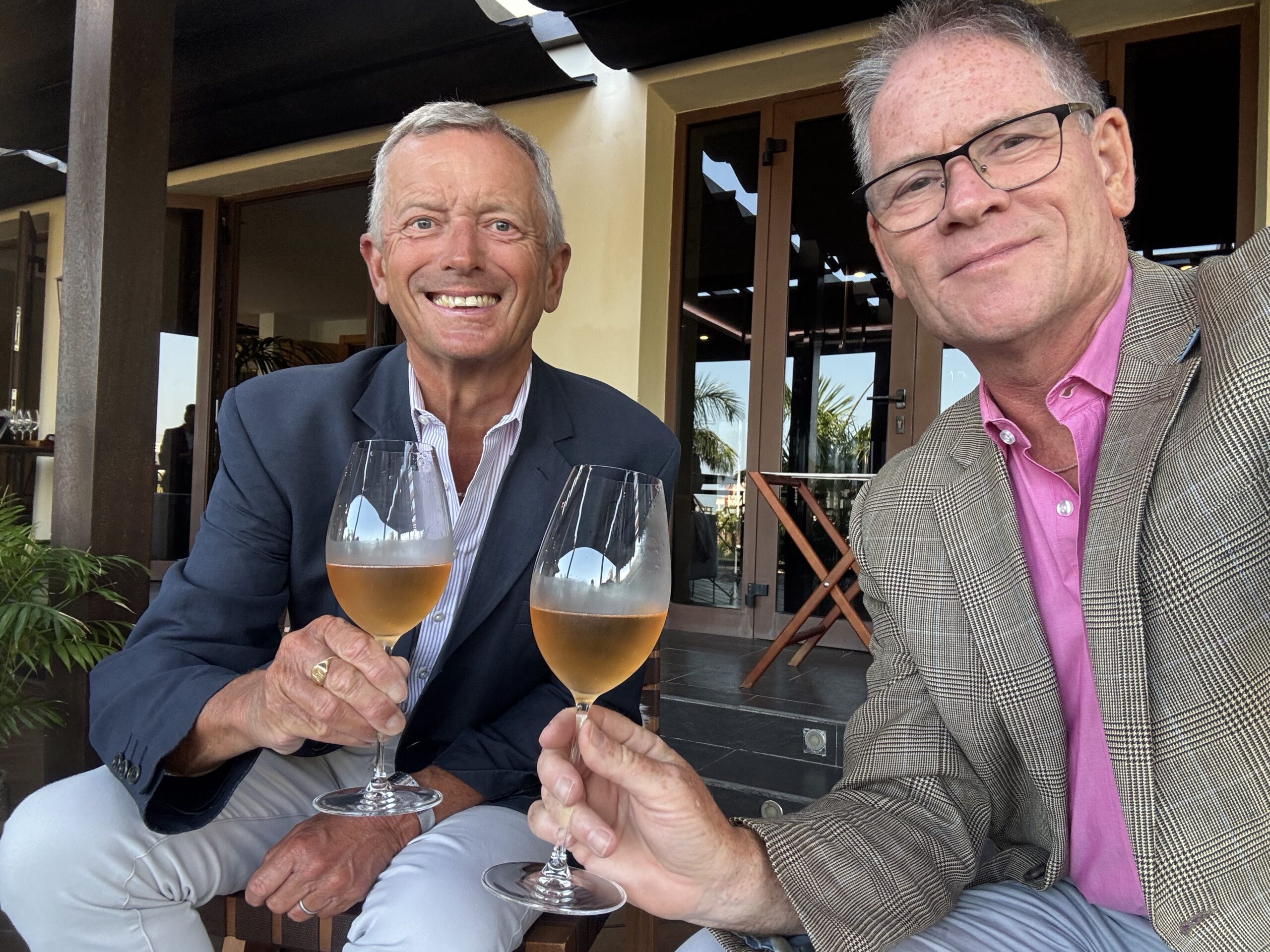Two men in blazers toast with wine glasses at an outdoor restaurant patio.