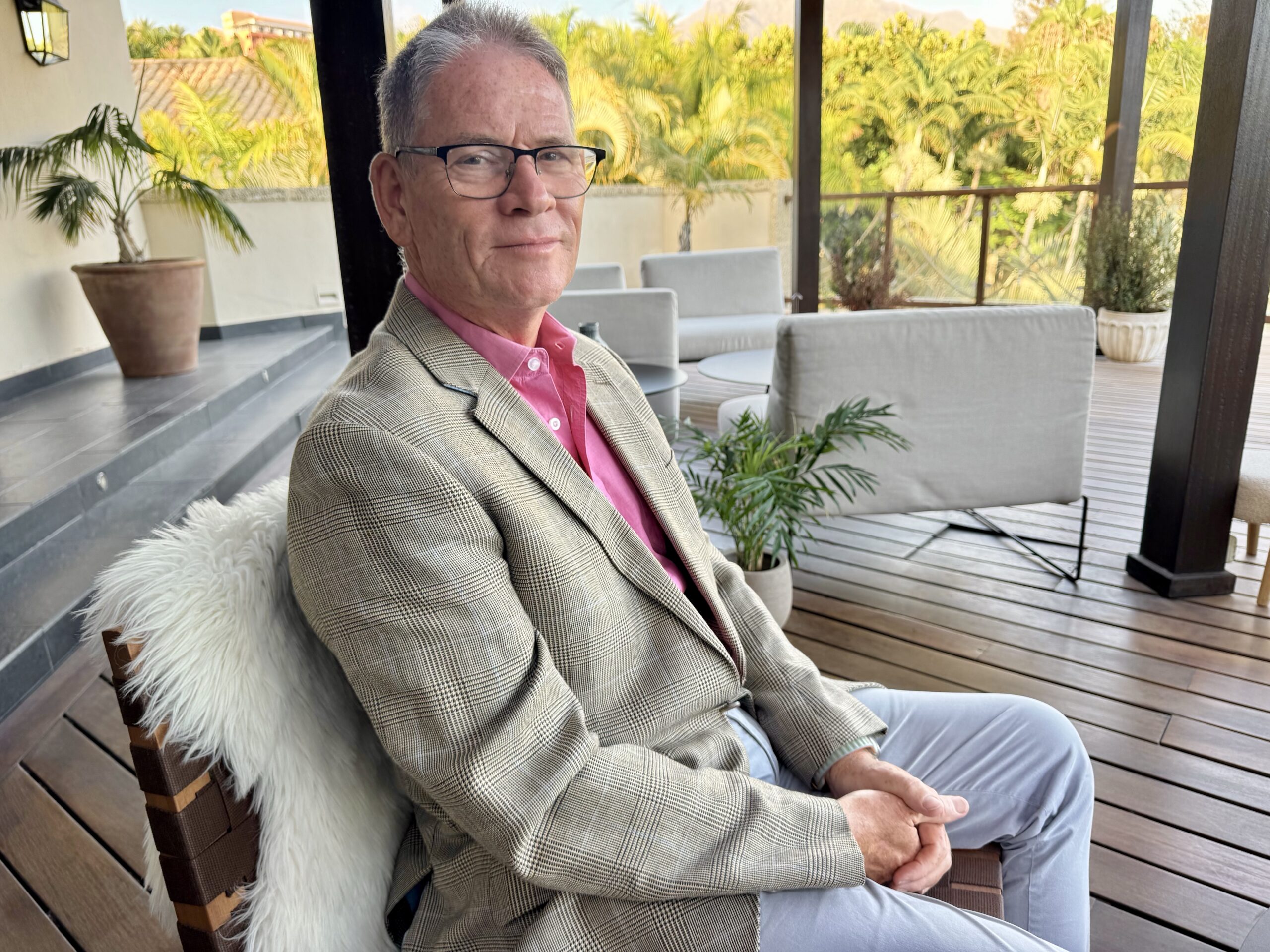 Older man in a beige plaid blazer and pink shirt sits on a wooden deck with potted plants and tropical greenery in the background.