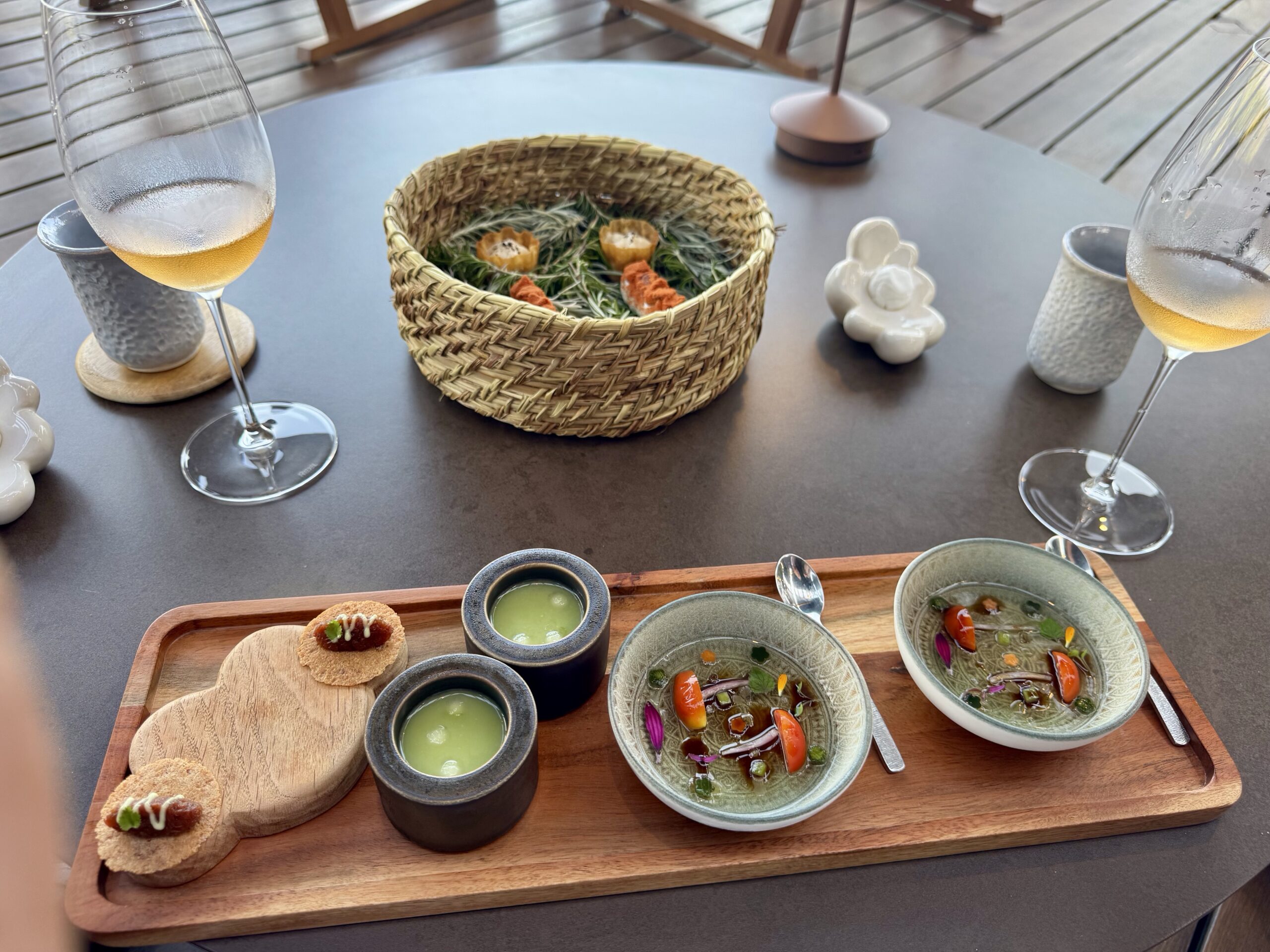 Table with a seafood-forward tasting menu: small bowls of green sauce and colorful plated items on a wooden board, two wine glasses nearby.