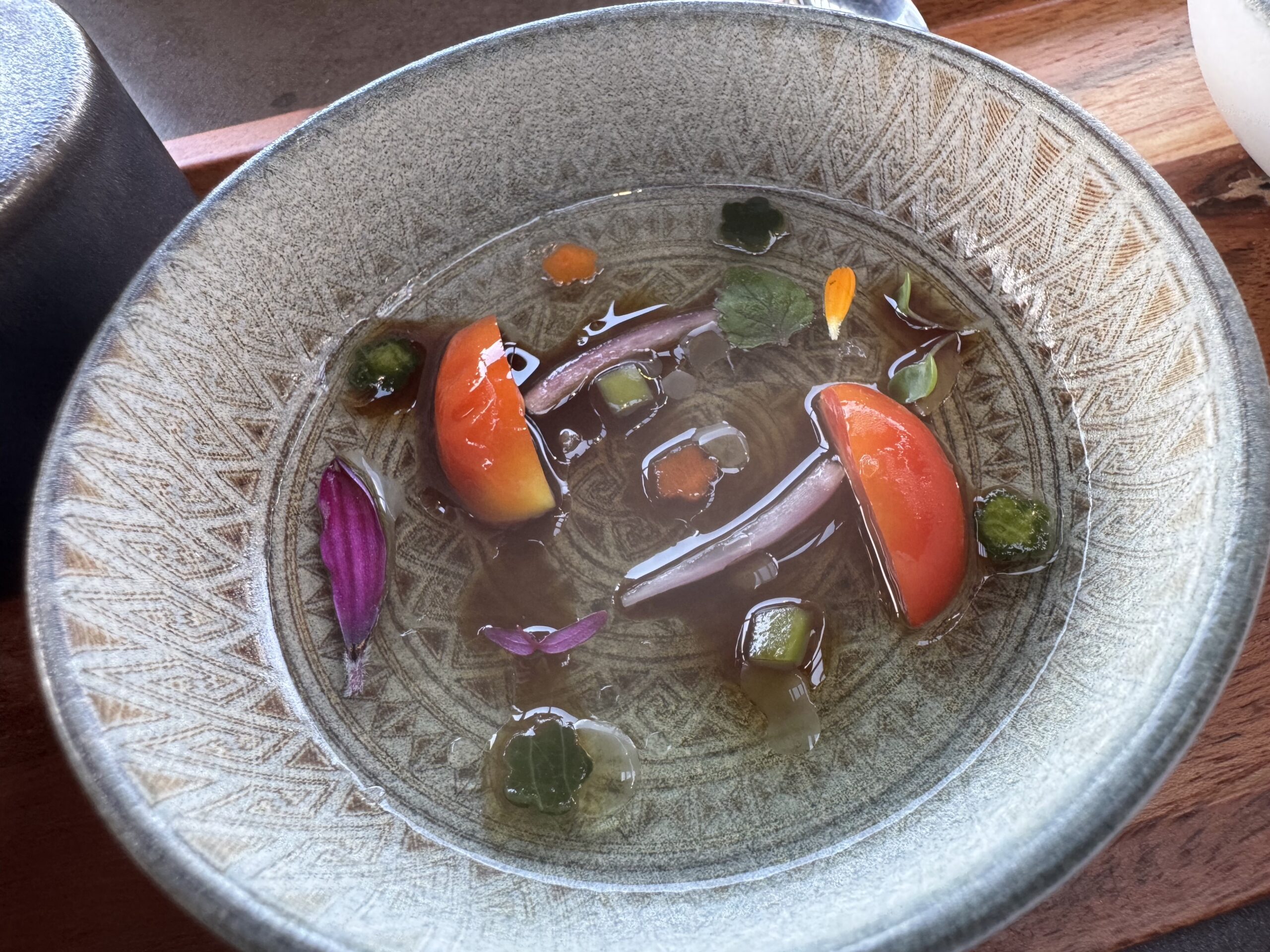 Close-up of a textured ceramic bowl with a light broth and floating colorful vegetables and petals (orange carrot, purple flower petals, green leaves)
